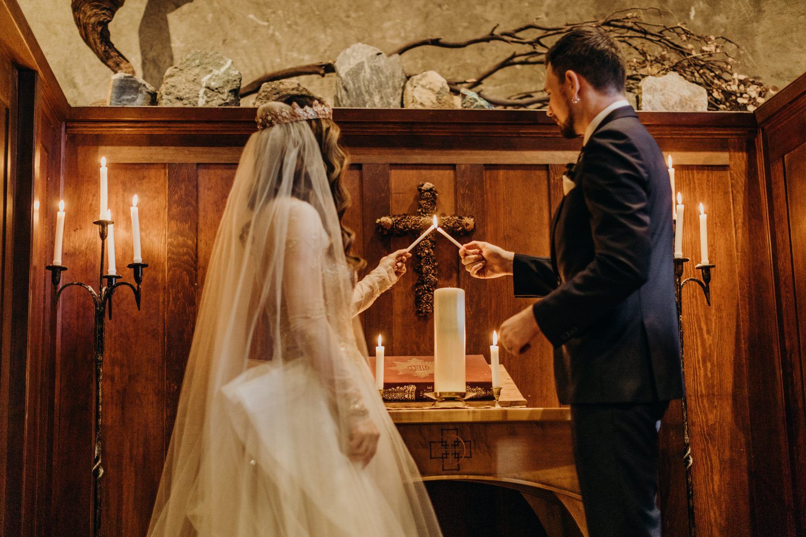The bride in her veil and the groom light a taper together from the two candles on either side of the altar, the tall unity candle unlit between them on an open book, a cross of pine cones on the wood panelled wall behind.