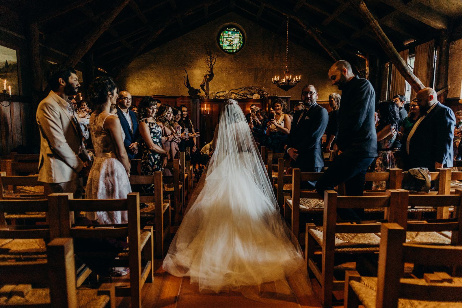 The bride from behind in her long tulle veil and skirt walking down the aisle of the church toward the stained glass window, guests standing in the wooden chairs on both sides, some smiling and holding up phones.