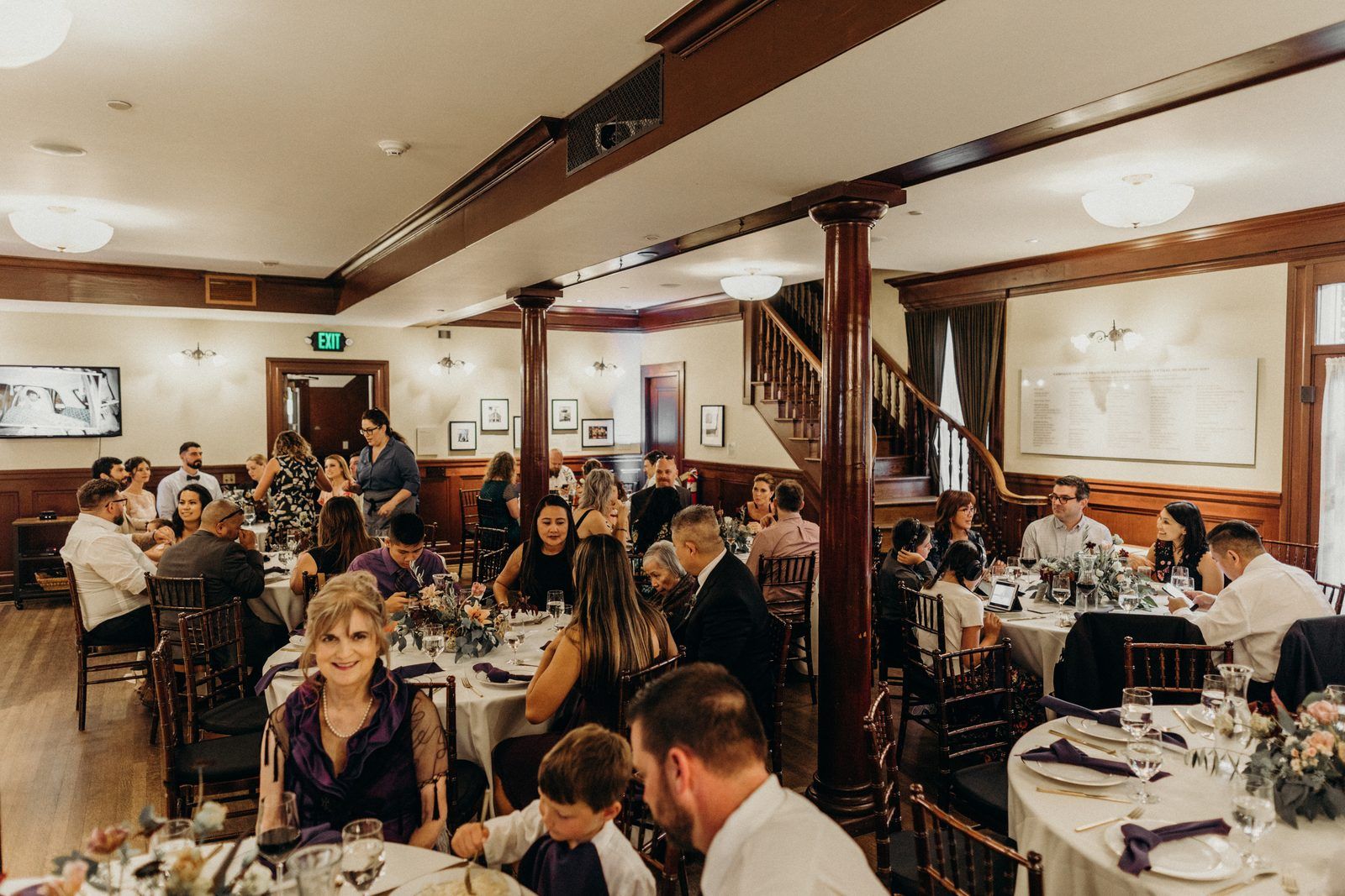 Guests seated at round tables in the ballroom during dinner, flowers and folded napkins on the tables and the wooden staircase behind.